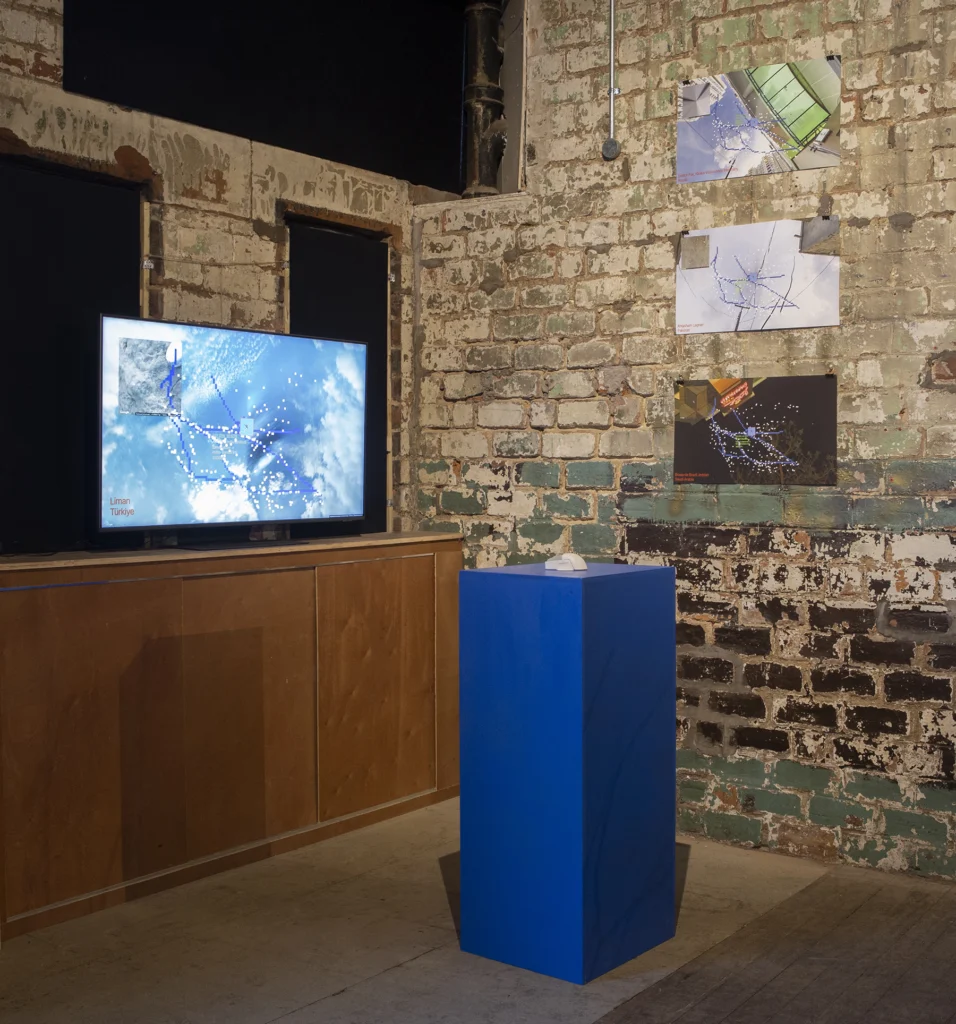 exhibition view of a sky on a large screen and blue plinth with prints on the wall in the background